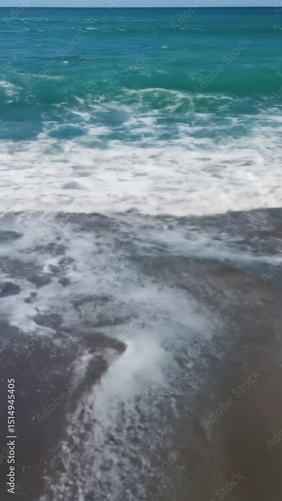 aerial close-up video of a vibrant turquoise ocean wave crashing onto a pristine sandy beach, churning into thick white foam creating intricate patterns on dark wet sand, illuminated by bright aerial close-up video of a vibrant turquoise ocean wave crashing onto a pristine sandy beach, churning into thick white foam creating intricate patterns on dark wet sand, illuminated by bright