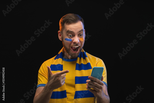 Studio shot of euphoric soccer supporter pointing at his phone screen with index finger celebrating triumph, his bet played, isolated over black background