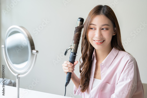 Fotografi Beautiful curly brunette, attractive asian young woman, girl smiling using curler, curling iron long hair with ceramic curler straightener at home