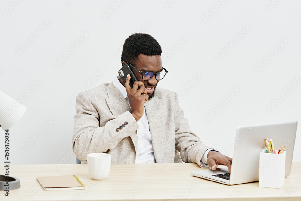 Professional man with dark skin, wearing glasses and beige blazer, talking on the phone while working on a laptop in a modern office space. Business, communication, technology, workspace, people Professional man with dark skin, wearing glasses and beige blazer, talking on the phone while working on a laptop in a modern office space. Business, communication, technology, workspace, people