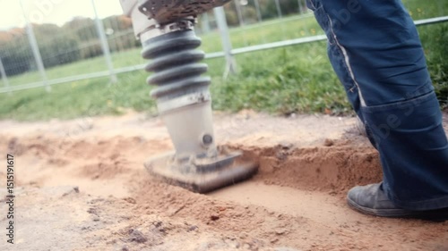 Close-up of a vibrating plate compactor being used by a worker to compact soil or sand at a construction site. The worker's legs in workwear are visible next to the equipment. Concept of groundwork