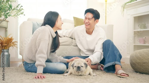 Happy Asian family couple enjoy and fun playing with senior Pug dog breed in living room. Husband and wife relaxing with domestic dog at home. Pet humanization, pet parent and senior pet healthcare.