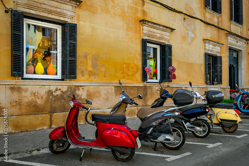scooters parked along the side of the road in front of a building with windows and shutters in Mahon, Spain.