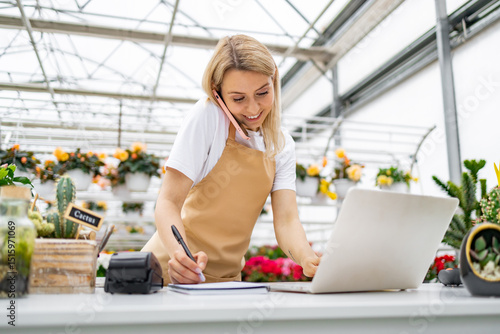 Obraz na płótnie A Caucasian woman working in a florist shop, taking a call and using a laptop while writing