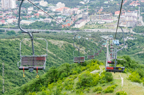 Chairlift suspended over green hillside in serene landscape. Empty chairs suggest quiet day. Dense vegetation covers slopes, contrasting with bright sky photo