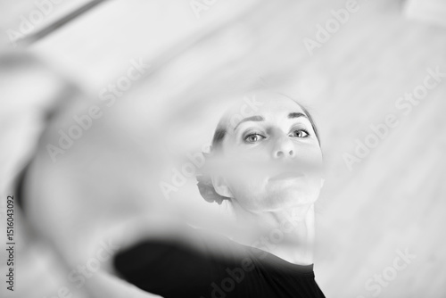 Young adult female ballerina with light hair captured reaching forward, creating dynamic motion. Black and white toning enhances focus on expression and poise, with blurred foreground