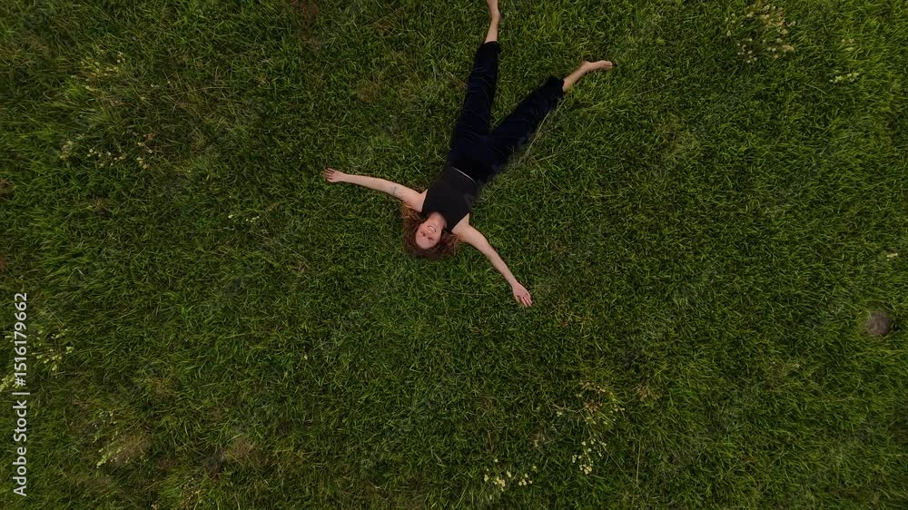 Barefoot woman lying on grass field and looking up to the sky. Aerial top down rising shot. Brunette girl relax in nature.