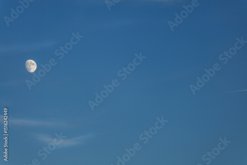 A waxing gibbous moon hangs in a clear blue sky with faint streaks of clouds. The stark contrast highlights the moon's craters against the vast expanse of a blue hour sky, exuding calmness.