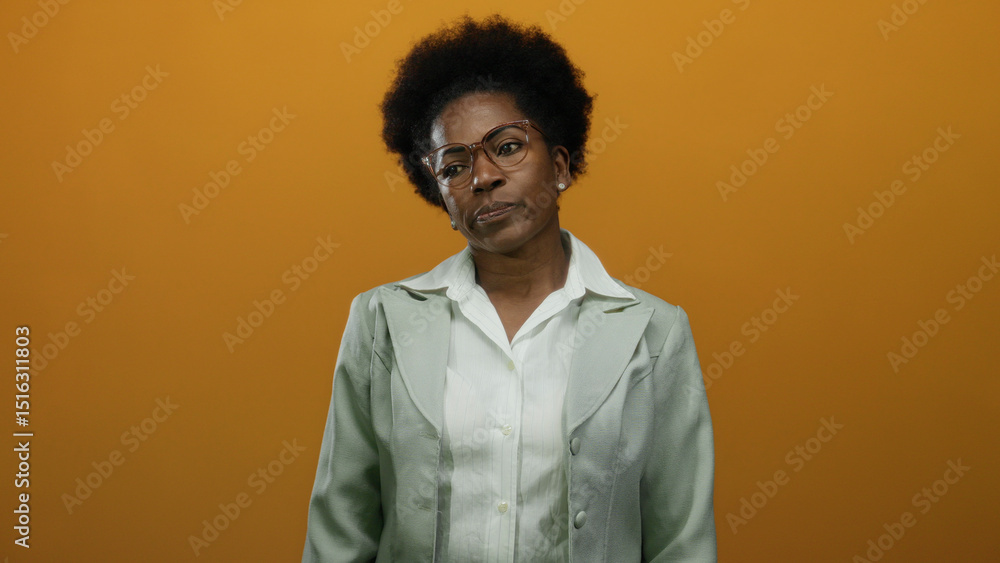 Obraz premium Woman with curly hair and glasses stands confidently against an isolated orange background, wearing a formal blazer, conveying professionalism and seriousness.