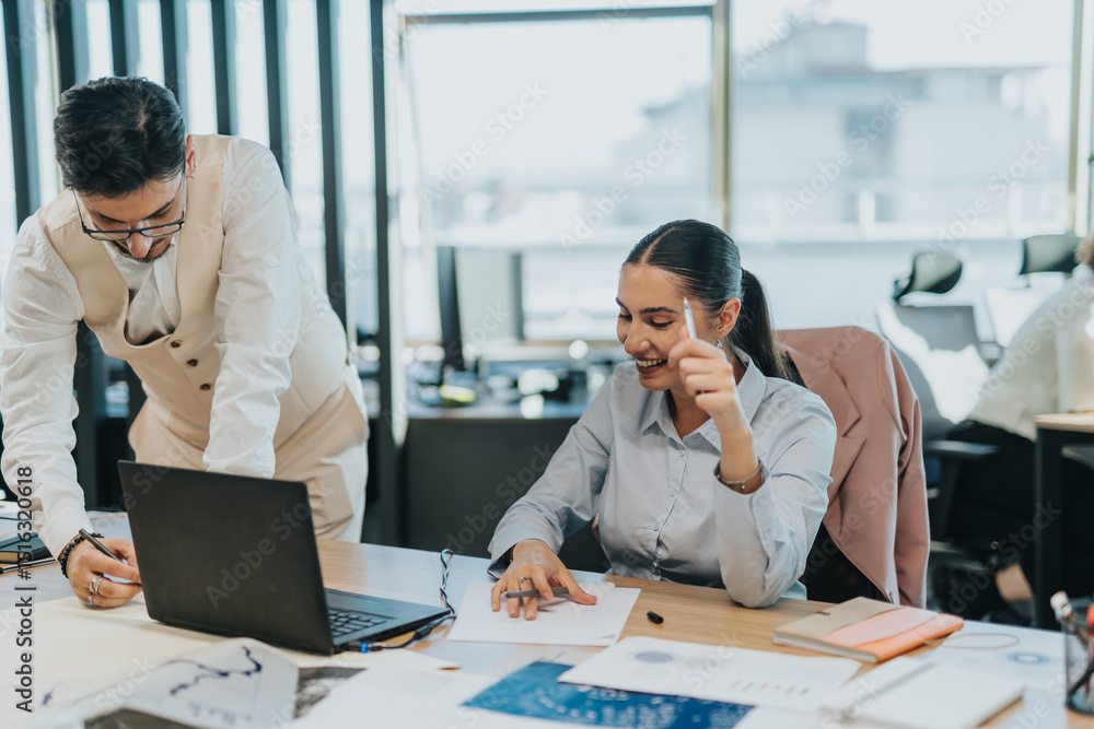 Fototapeta premium Two business employees working together in a modern office setting, discussing ideas and analyzing data on a laptop, fostering teamwork and professional collaboration.