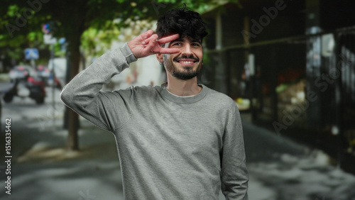 Young man smiling and making a peace sign outdoors on a sunny urban street under the shade of trees, dressed casually in a grey sweatshirt, portraying a relaxed and confident demeanor.