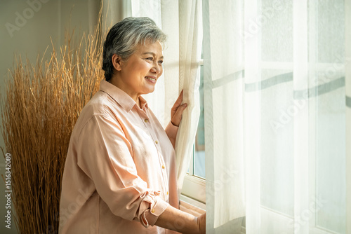 Happy senior Asian woman looking out window with morning sunlight and peaceful smile