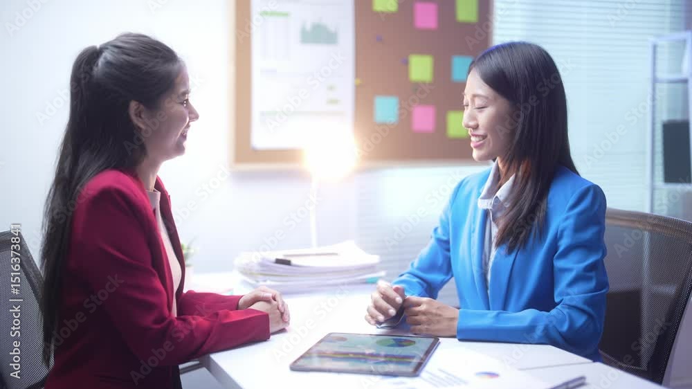 Two women are sitting at a desk, one of them is wearing a red jacket. They are talking to each other Two women are sitting at a desk, one of them is wearing a red jacket. They are talking to each other