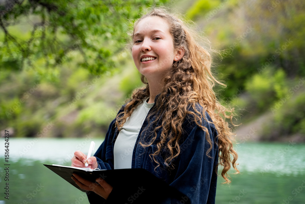 Naklejka premium Portrait of young woman with curly hair smiling at camera writing in notebook with pen taking notes journaling keeping diary dressed in blue shirt standing in mountain forest near lake outdoors