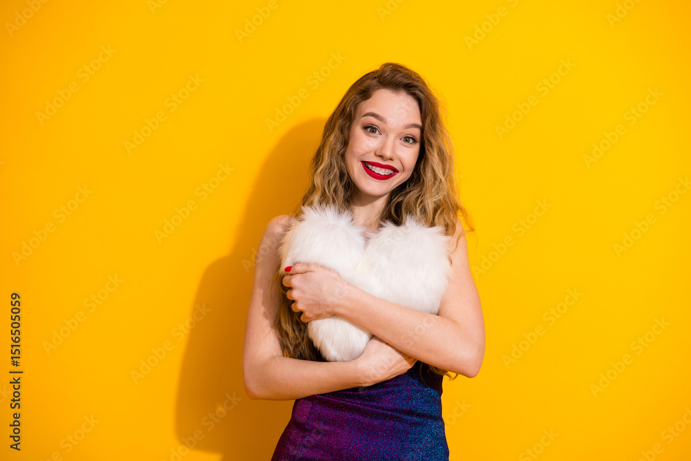 Fototapeta premium Cheerful young woman in fashionable dress holding a soft heart pillow against a bright yellow background, smiling warmly