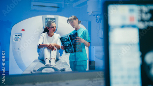 Charming woman sitting on CT scanner bed discussing scan results with female doctor in turquoise uniform. Medical specialist holding radiographic images explaining diagnosis after successful procedure