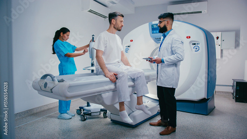 Attractive male sitting upright on MRI bed having discussion with doctor holding digital tablet. Smiling nurse adjusting nearby device while preparing for procedure in clean clinical environment.
