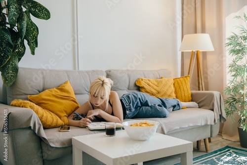 Εκτύπωση καμβά Young woman relaxing on sofa, drawing in notebook at home