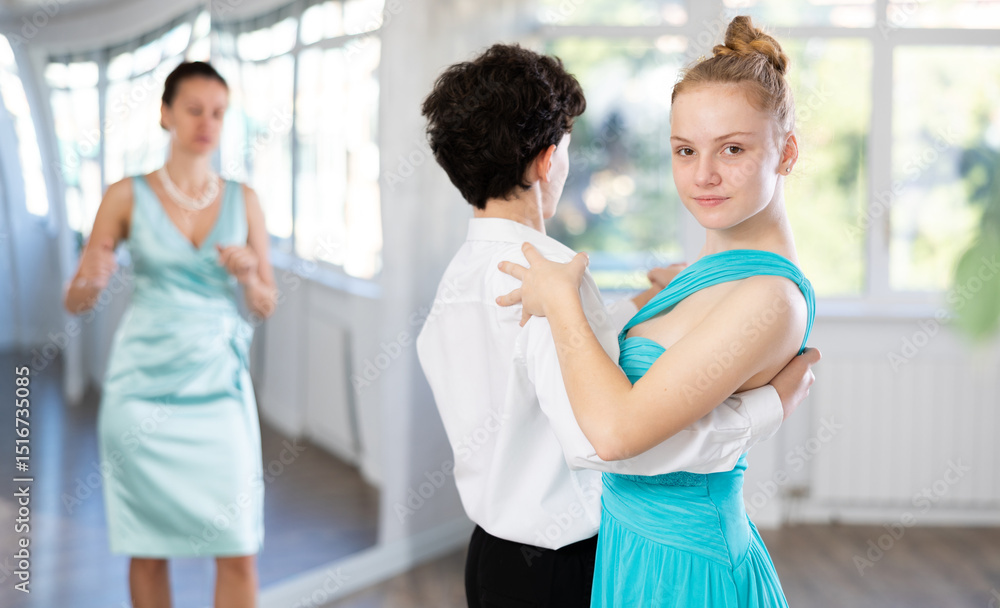 Obraz premium Motivated adolescent ballroom dancers, girl and boy in performance outfit practicing elegant dance moves in pair in bright studio, with female coach supervising in background