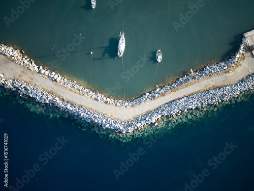 Aerial view of a tranquil Greek breakwater stretching into deep blue waters, with moored sailboats casting shadows on the calm sea — captured at golden hour in Agii Apostoli