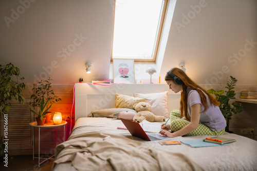 A young woman uses a laptop and writes in a notebook while sitting on her bed at home. Headphones on her head.