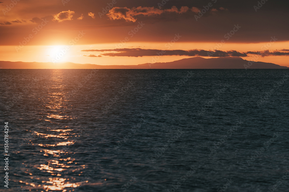Obraz premium Golden sunset reflecting on calm sea with distant mountains and clouds creating a dramatic and peaceful atmosphere