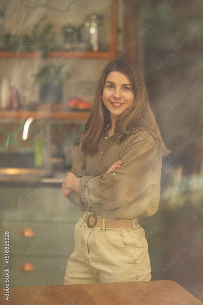 Fototapeta premium Light-Skinned European Woman Standing with Arms Crossed and Smiling in Cozy Kitchen