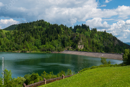 Fototapeta Naklejka Na Ścianę i Meble -  Wide view of Czorsztyn Lake dam in Poland with surrounding green forest and hills. Concrete slope structure meets calm water under partly cloudy sky.
