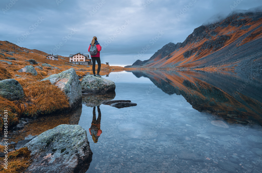 Fototapeta premium Młoda kobieta wędrująca z plecakiem na skale jesienią. Dziewczyna i zapierający dech w piersiach widok na górskie jezioro odbijające okoliczne szczyty, pomarańczową trawę i pochmurne, dramatyczne niebo. Szwajcaria jesienią.