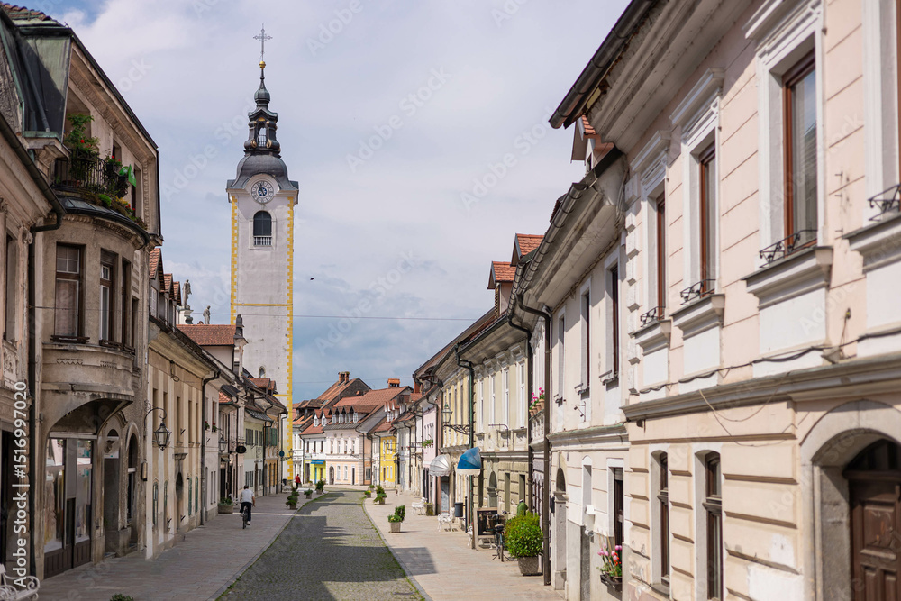 Fototapeta premium Charming old town street in Kamnik, Slovenia, featuring cobblestone pavement, historic pastel buildings, and the iconic church tower. Locals stroll peacefully under a partly cloudy sky.