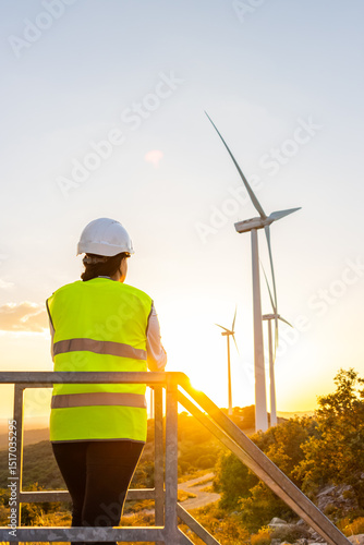 Rear view of female engineer on metal platform near wind turbines at sunset