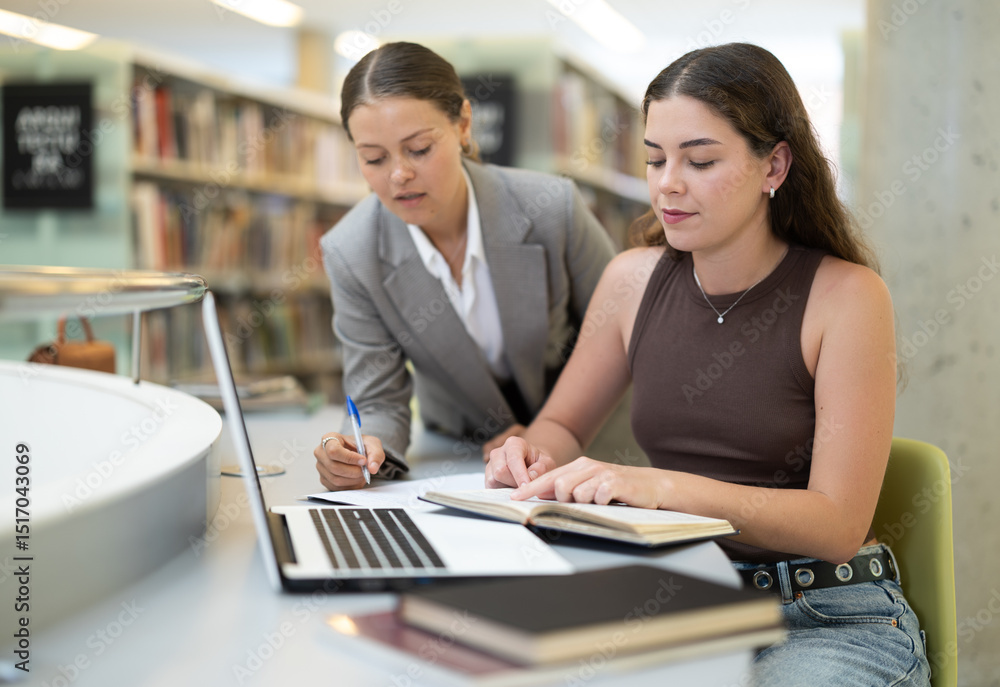 Focused female student works at a notebook in a library and makes notes in a notebook, next to her is a classmate who helps her. Students come to the library and study together Focused female student works at a notebook in a library and makes notes in a notebook, next to her is a classmate who helps her. Students come to the library and study together