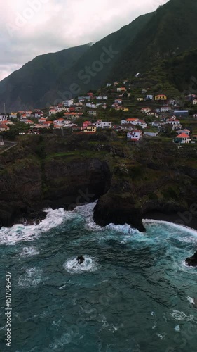 Cinematic aerial footage of a small coastal town perched on dramatic rocky cliffs along the Atlantic Ocean on Madeira Island, Portugal.