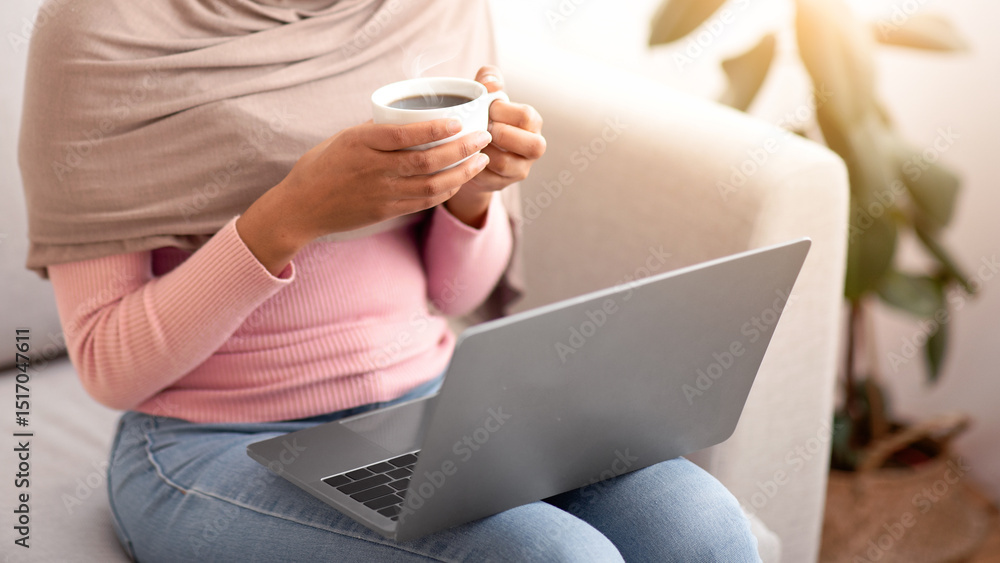 Naklejka premium Online work, education, communication. Unrecognizable black woman in hijab using laptop and drinking coffee at home. Young Muslim lady having web lesson or conference indoors