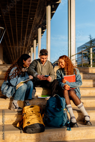 Three cheerful university students are sitting on campus stairs, sharing content on a smartphone, enjoying a break between classes