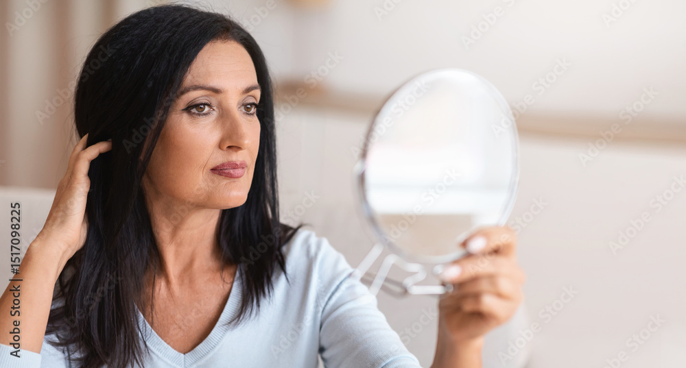 Portrait of beautiful middle-aged brunette lady holding mirror and looking at her reflection, touching her hair with hand. Confident adult woman thinking about skin care and body treatment Portrait of beautiful middle-aged brunette lady holding mirror and looking at her reflection, touching her hair with hand. Confident adult woman thinking about skin care and body treatment