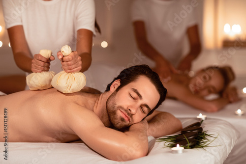 Couple Relaxing During Herbal Back Massage With Aromatic Bags And Aromatherapy Lying On Beds Indoors, With Eyes Closed. Body Care And Exotic Relaxation Concept. Selective Focus