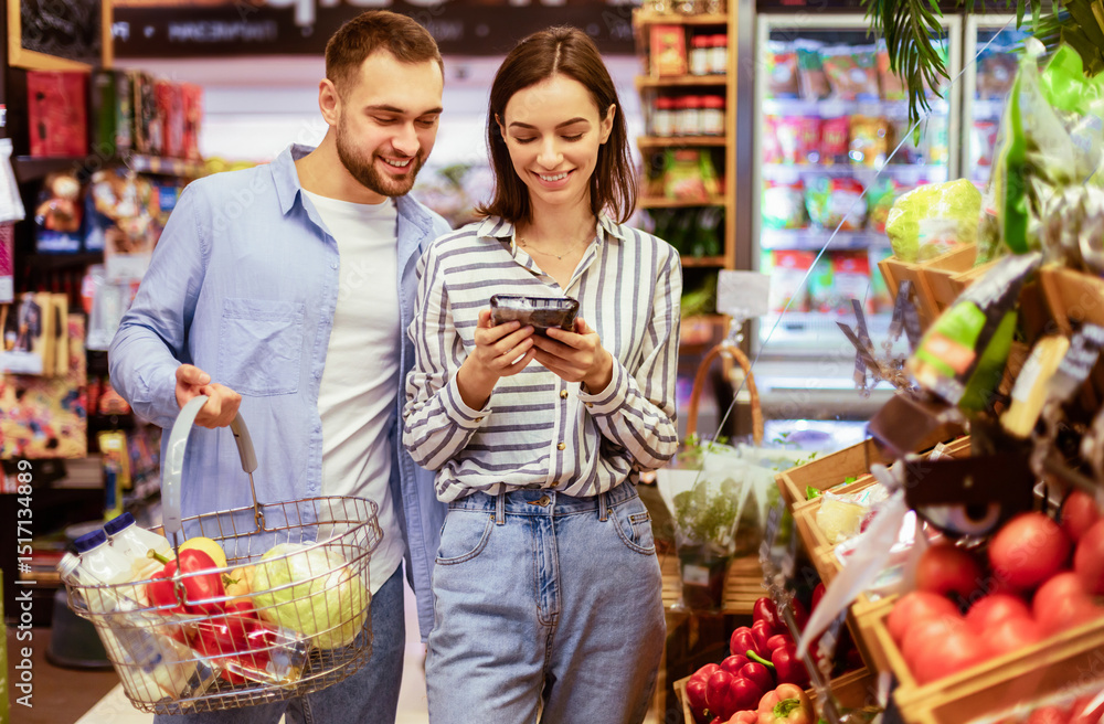 Obraz premium Smiling Millennial Couple Shopping Groceries In The Hypermarket, Two Happy Consumers Buying Food Walking With Cart In Wholesale Shop, Choosing Eco Vegetables And Fruits, Checking Label And Expiry Date