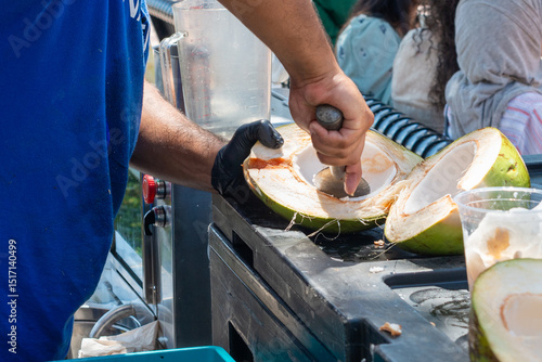A man wearing a blue shirt holds a large knife, cleans a fresh coconut at a farmer's market. The fruit is used to make an organic tropical coconut fruit drink for sale at a street vendor's cart.
