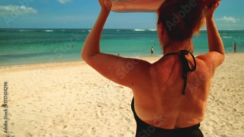 Old woman carries surfboard over head in hands. Senior female in swimsuit goes surfing on waves. Elderly lady beginner student goes on surfing session at tropical location at sunrise. Above head