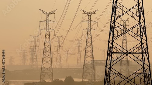 Power transmission towers and high-voltage lines under sunrise silhouette, dramatic long-exposure photography with warm golden tones, aerial view of energy infrastructure in natural landscape