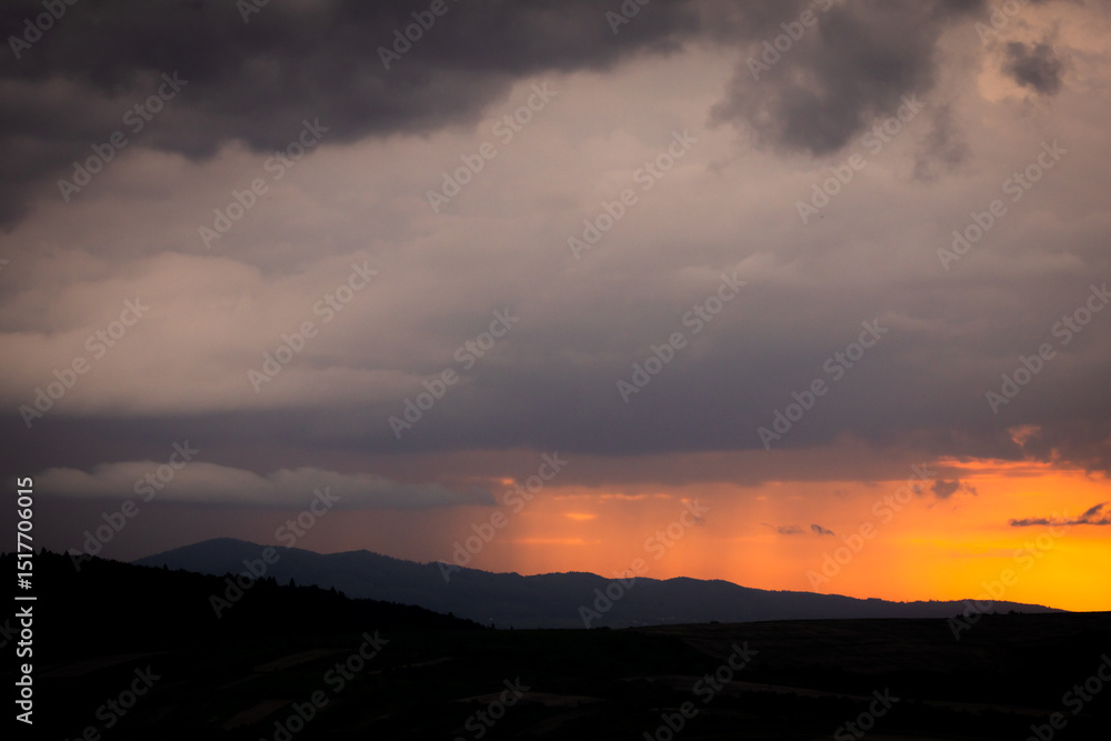 Obraz premium Vibrant orange sunset behind a silhouette of leafy trees under heavy clouds, capturing the beauty and contrast of nature in a summer evening storm.