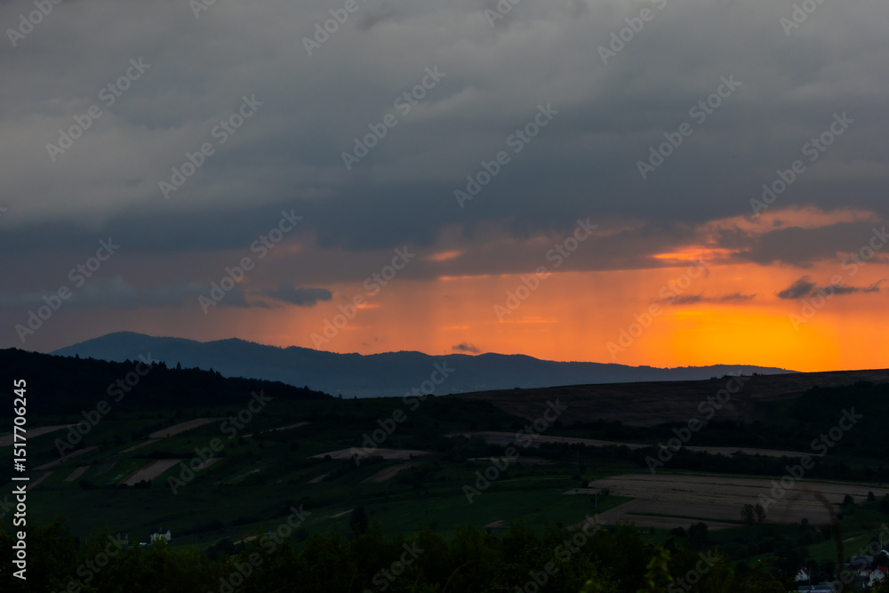 Fototapeta premium Vibrant sunset light pierces through layers of clouds over farmland and hills, casting long shadows and painting the sky with warm tones. A captivating rural moment.
