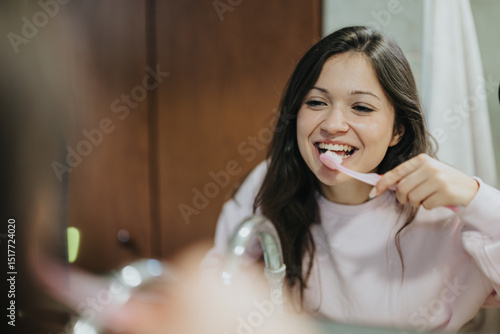 A cheerful young woman in a pastel-colored top brushing her teeth while standing before a bathroom mirror, promoting daily dental health practices and hygienic routines in a congenial setting.
