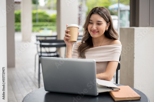Long hair asian woman is smiling as she sitting holding coffee and looking at laptop on cafe table. Free Download Long hair asian woman is smiling as she sitting holding coffee and looking at laptop on cafe table. - GFXLoad