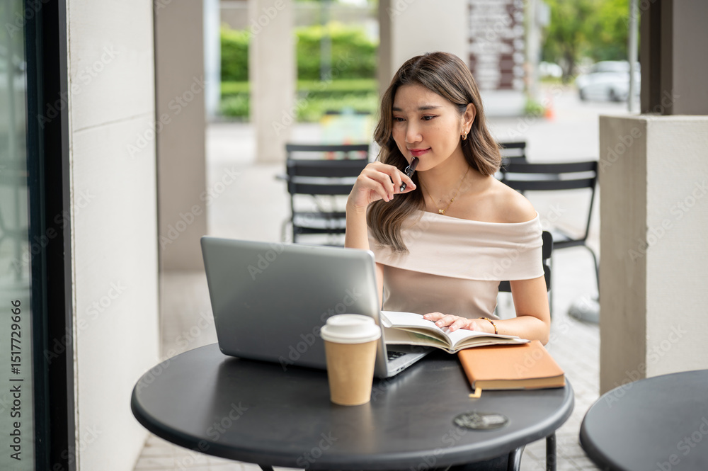 Asian woman looking at laptop with pen touching lip and hand on book with coffee aside at cafe table Asian woman looking at laptop with pen touching lip and hand on book with coffee aside at cafe table