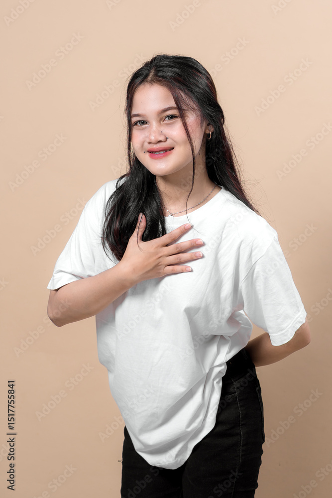 Young Asian woman in a white t-shirt and black pants smiles warmly while placing one hand on her chest, conveying sincerity, appreciation, and a friendly, humble attitude. Young Asian woman in a white t-shirt and black pants smiles warmly while placing one hand on her chest, conveying sincerity, appreciation, and a friendly, humble attitude.