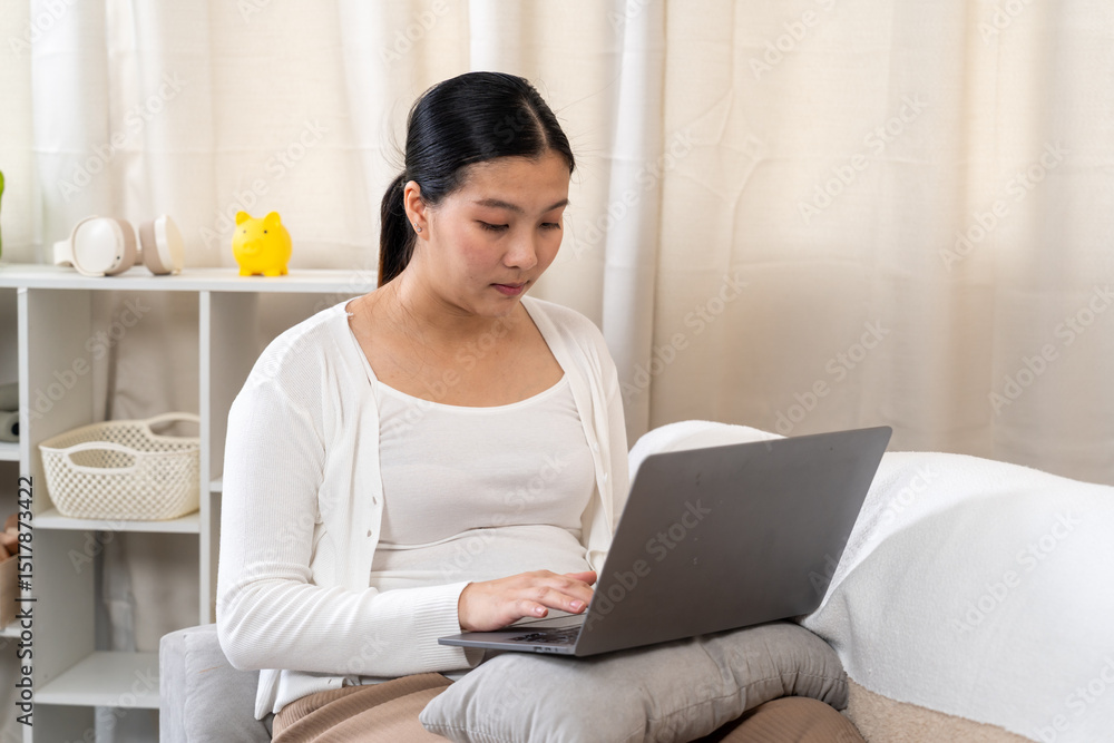 Pregnant Asian woman working on laptop while sitting on sofa with pillow support. Wearing white cardigan and brown pants. Focused expression shows commitment to productivity, comfort, and self-care Pregnant Asian woman working on laptop while sitting on sofa with pillow support. Wearing white cardigan and brown pants. Focused expression shows commitment to productivity, comfort, and self-care