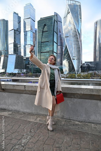 A young woman of European appearance takes a selfie with a smartphone against the backdrop of Moscow City skyscrapers outdoors