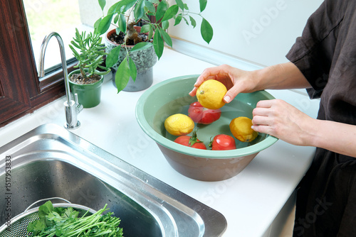 A woman washes fresh lemons, tomatoes, and leafy greens in a kitchen sink, preparing healthy ingredients near a sunlit window.